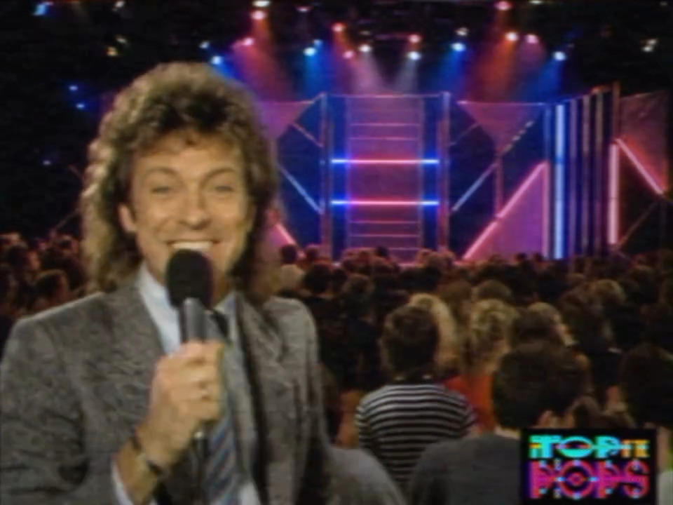 A frame from CIAO64's 1980s Top of the Pops-style reel made with AnalogTV: a presenter holding a microphone in front of a neon-lit stage set and crowd, rendered with visible scanlines and a soft CRT glow